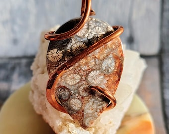May include: A pendant featuring an oval-shaped piece of fossilized coral with a distinct floral pattern in shades of brown, gray, and cream. The stone is wrapped in polished copper wire that forms a bail at the top for hanging.