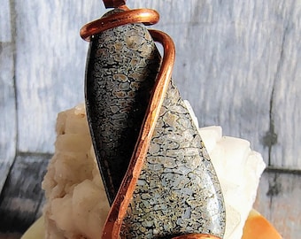 May include: A teardrop-shaped pendant featuring a dark gray and tan mottled stone encased in a polished copper wire wrap. The pendant includes a copper bail at the top for suspension, showcasing a smooth, polished surface texture.