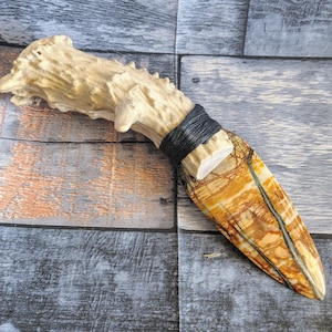 May include: A handcrafted knife with a natural antler handle and a blade featuring a brown, tan, and black patterned stone. The handle is wrapped with black material, creating a rustic and unique aesthetic.