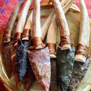 May include: A collection of six arrowheads with different colored stones, each attached to a brown antler handle with a brown leather wrap.