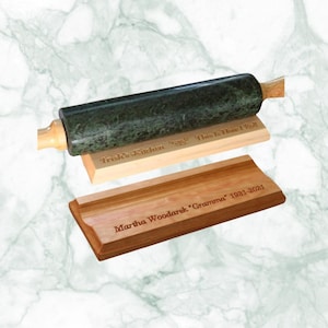Può includere: Un mattarello in marmo verde con manici in legno poggia su una base in legno. Sotto, due assi di legno incise con testo. Una recita "Trish's Kitchen" e l'altra "Martha Woodarek 'Gramma' 1931-2021".