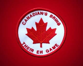 May include: A circular embroidered patch featuring a white background with a red maple leaf in the center. The text CANADIAN'S BRING THEIR EH GAME is stitched in red capital letters around the perimeter of the patch, which has a red border.
