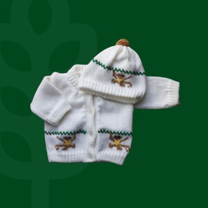May include: A white knitted long-sleeve cardigan and matching beanie hat set. The cardigan features a button-front closure and a horizontal green stitched pattern with small brown and yellow monkey motifs. The beanie has a brown pom-pom on top.