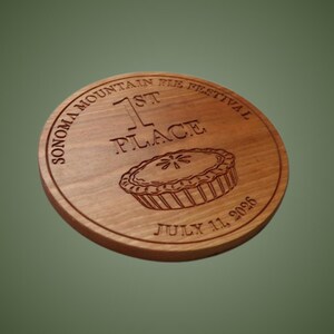 Könnte beinhalten: Eine kreisförmige Holzplakette mit einer Gravur. Der Text lautet Sonoma Mountain Pie Festival, 1st Place und July 11, 2026. Eine Grafik eines Kuchens ist auf der Holzoberfläche zentriert, die eine natürliche Maserung und einen warmen Braunton aufweist.
