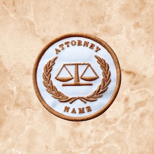 May include: A circular embroidered patch with a white background and a brown border. The design features a brown scale of justice icon centered within a brown laurel wreath. The text "ATTORNEY" is arched above and "NAME" is below the icon.