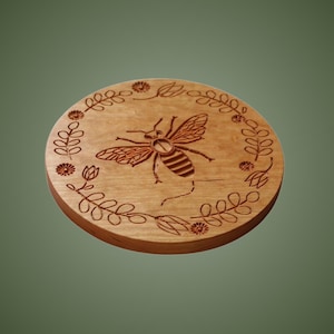May include: A circular wooden coaster featuring a laser-engraved design of a bee in the centre, surrounded by a decorative border of small flowers and leafy vines. The natural light brown wood grain is visible across the flat, round surface.