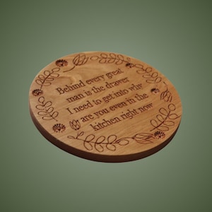 May include: A round wooden coaster featuring a laser-engraved floral border and the text: Behind every great man is the drawer I need to get into why are you even in the kitchen right now. The natural wood grain is visible across the surface.