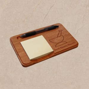 May include: A rectangular wooden desk organiser featuring a carved groove for a black ballpoint pen, a square stack of light yellow sticky notes, and an engraved mortar and pestle icon with the text Sarah Nguyen, PharmD on the surface.