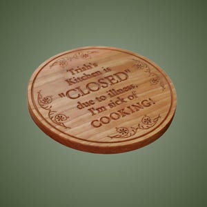 May include: A round wooden chopping board with a natural light brown grain. The surface features laser-engraved text that reads: Trish's Kitchen is "CLOSED" due to illness. I'm sick of COOKING! Decorative floral scrollwork accents the top and bottom.
