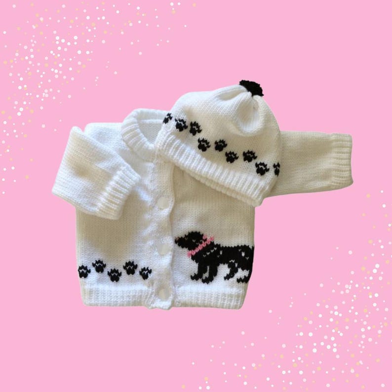 May include: A white knitted baby cardigan and matching hat set. The cardigan features a black dog design with a pink collar and paw prints. The hat has paw prints and a black pom-pom. The set is displayed on a pink background.