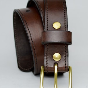 May include: A brown leather belt with a brass buckle. The belt has a stitched edge and two brass rivets.