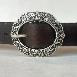 May include: A dark brown leather belt featuring an oval-shaped silver-toned metal buckle. The buckle has an intricate, embossed floral and scrollwork pattern along its outer edge and includes a standard prong for securing the belt strap.
