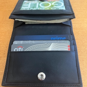 May include: A black leather wallet featuring a clear ID window containing a card with the text GOLF, multiple interior card slots holding a blue Prime card and a grey Citi card, and a silver metal snap button closure at the bottom.