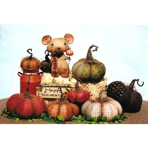 May include: A collection of handmade patchwork pumpkins in various colors and patterns, including red, green, brown, and beige. A small mouse doll with a red scarf is holding a miniature pumpkin. The pumpkins are arranged on a bed of straw with a sign that reads "Happy Harvest".