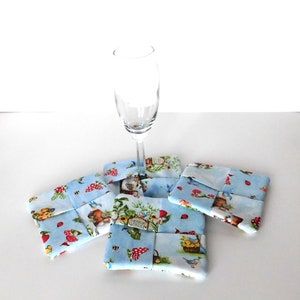May include: Four square fabric coasters with a light blue background and a whimsical garden print featuring mushrooms, birds, and gnomes. A champagne flute rests on top of the coasters. The coasters are approximately 4 inches square.