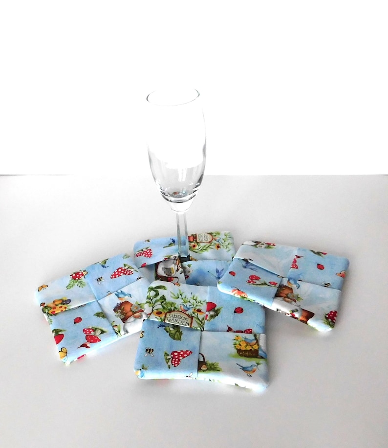 May include: Four square fabric coasters with a light blue background and a whimsical garden print featuring mushrooms, birds, and gnomes. A champagne flute rests on top of the coasters. The coasters are approximately 4 inches square.