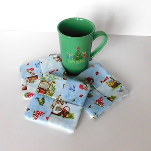 May include: A green mug with a Christmas tree design and the words "Happy Holidays" sits atop a set of fabric coasters. The coasters feature a light blue background with a whimsical garden print, including gnomes, mushrooms, and dragonflies.