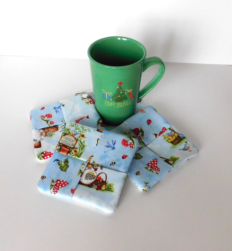 May include: A green mug with a Christmas tree design and the words "Happy Holidays" sits atop a set of fabric coasters. The coasters feature a light blue background with a whimsical garden print, including gnomes, mushrooms, and dragonflies.