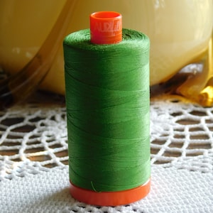 May include: A spool of green thread with an orange plastic top. The spool is sitting on a white crocheted doily.