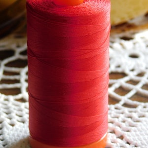 May include: A spool of bright red thread with an orange plastic top and bottom. The spool is sitting on a white crocheted doily.