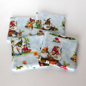 May include: Four square fabric coasters with a light blue background featuring whimsical garden gnomes, mushrooms, flowers, and butterflies. The gnomes wear red, green, and yellow hats. The coasters are a charming addition to any home decor.