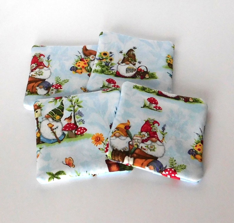 May include: Four square fabric coasters with a light blue background featuring whimsical garden gnomes, mushrooms, flowers, and butterflies. The gnomes wear red, green, and yellow hats. The coasters are a charming addition to any home decor.