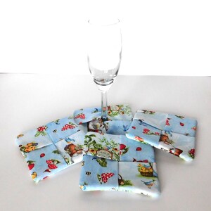 May include: A clear champagne flute rests on four square fabric coasters. The coasters feature a light blue background with a whimsical print of mushrooms, gnomes, and garden elements. The coasters are approximately 4 inches square.