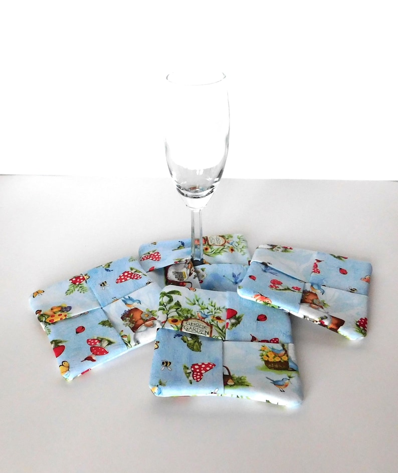 May include: A clear champagne flute rests on four square fabric coasters. The coasters feature a light blue background with a whimsical print of mushrooms, gnomes, and garden elements. The coasters are approximately 4 inches square.