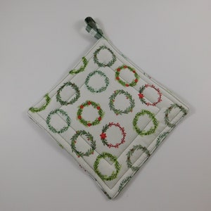 May include: A white quilted pot holder with a green and white checkered hanging loop. The pot holder is decorated with a repeating pattern of green wreaths with red berries.