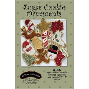 May include: A collection of felt ornaments shaped like Christmas cookies, including a snowman, a reindeer, a candy cane, a stocking, a mitten, a bell, a star, a poinsettia, a tree, a gingerbread man, and Santa Claus. The ornaments are on a white plate and the text "Sugar Cookie Ornaments #1822 Finished sizes: vary approximately 3" - 5" ©2018" is printed below the plate. The text "All Through the Night Folk Art designs with a bit of Whimsy by Bonnie Sullivan Made in the USA" is printed in a circle on the left side of the image.
