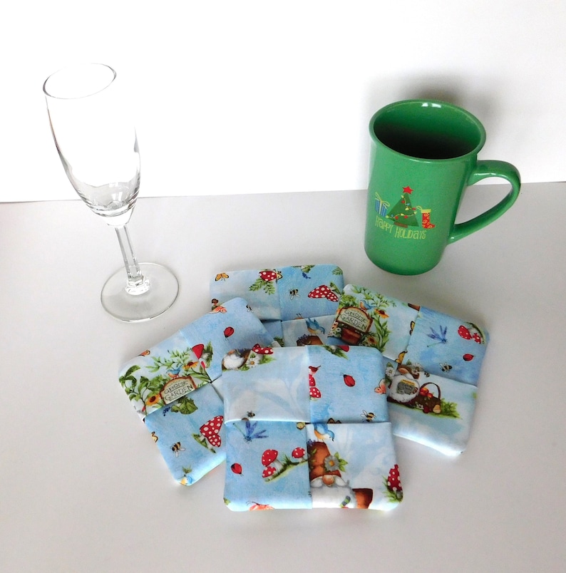 May include: Four square fabric coasters with a whimsical garden scene on a light blue background. A green mug with a Christmas tree design and a champagne flute are also present. The coasters feature mushrooms, bees, and garden gnomes.
