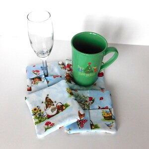 May include: A set of four square fabric coasters with a light blue background and a whimsical gnome and garden scene. A green mug with "Happy Holidays" text and a champagne flute rest on the coasters. The coasters are approximately 4 inches square.