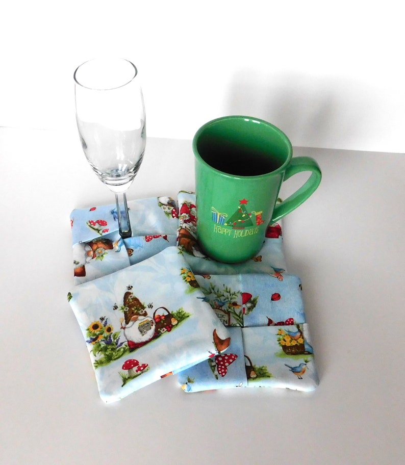 May include: A set of four square fabric coasters with a light blue background and a whimsical gnome and garden scene. A green mug with "Happy Holidays" text and a champagne flute rest on the coasters. The coasters are approximately 4 inches square.