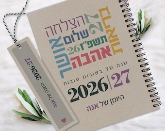 Może przedstawiać: Planer spiralny z beżową okładką zawierającą hebrajski tekst w różnych kolorach oraz lata 2026-2027. Dołączona jest pasująca zakładka przymocowana czarno-białym sznurkiem, na której również widnieją lata 2026-2027 oraz hebrajski tekst.