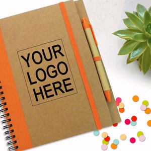 Könnte beinhalten: Ein braunes Spiralnotizbuch mit orangefarbenem Rücken und einem passenden Gummibandverschluss. Das Notizbuch hat ein weißes Etikett mit dem Text "YOUR LOGO HERE" in Schwarz gedruckt. Ein beiger Stift mit orangefarbener Spitze liegt auf dem Notizbuch.