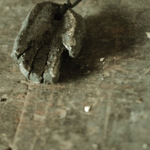 Puede incluir: Un colgante con forma de mano hecho de arcilla gris oscuro que cuelga de un cordón negro. El colgante tiene una superficie rugosa y texturizada.