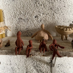May include: Five small clay figurines, each with a human-like form, are arranged on a wooden shelf. The figurines are all brown and have simple features. One figurine is sitting upright, while the other four are sitting with their legs crossed. The figurines are all facing forward.