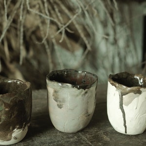 May include: Three small ceramic cups with a rustic, hand-crafted look. The cups are glazed in shades of brown, white, and grey, with a slightly uneven finish.