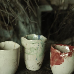 May include: Three handmade ceramic cups with uneven rims. The cups are off-white with splashes of blue, green, and red. The cups are displayed on a dark wooden surface.