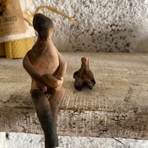 May include: Two small clay figurines, one sitting with its legs crossed and the other standing with its arms outstretched, are displayed on a wooden shelf. The figurines are a light brown colour and have a rustic, handmade appearance.