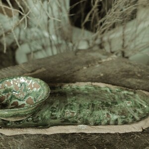 May include: A green ceramic bowl with brown speckles sits on a green ceramic plate with a rough, uneven edge. Both pieces are handmade and have a rustic, earthy feel.