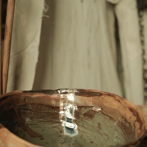 May include: A brown ceramic bowl with a green glaze and a white stripe around the rim. The bowl is filled with water and reflects the surrounding environment.
