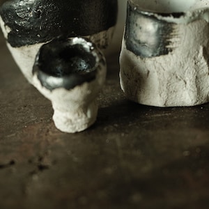 May include: Three ceramic cups with a white glaze and black accents. The cups are sitting on a wooden surface.