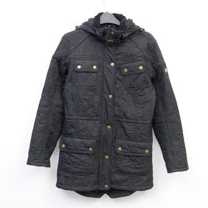 Puede incluir: Chaqueta acolchada negra con capucha, cuatro bolsillos delanteros y botones dorados. La chaqueta está colgada de una percha sobre un fondo blanco. La chaqueta tiene un patrón de diamantes.