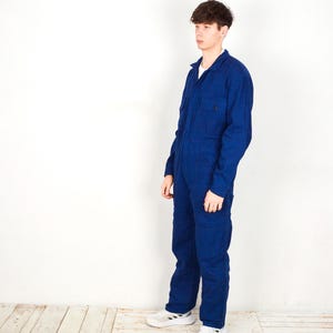 May include: A dark blue jumpsuit with long sleeves and a collared neckline. The workwear-style jumpsuit features multiple pockets and a button-up front. The model is wearing white sneakers. The jumpsuit is made of a durable fabric.