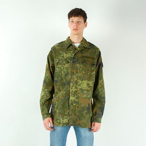 Può includere: Una camicia mimetica a maniche lunghe nei toni del verde, marrone e nero. La camicia ha una chiusura con bottoni, due tasche sul petto e spalline sulle spalle. La persona indossa jeans blu.