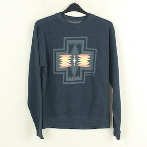 Puede incluir: Una sudadera azul oscuro de manga larga con un diseño geométrico en la parte delantera. El diseño es una forma de cruz con un patrón de naranja, amarillo y gris. La sudadera está colgada en una percha negra.