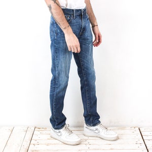 May include: Blue denim jeans with a classic five-pocket design. The jeans have a medium wash with subtle fading and whiskering details. The model is wearing white trainers. The jeans are a straight-leg fit.