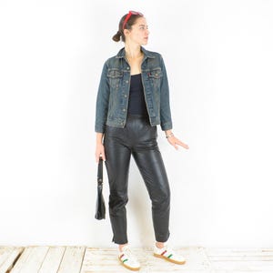 Puede incluir: Una mujer con una chaqueta vaquera azul, pantalones de cuero negros y zapatillas blancas con rayas verdes y rojas. Sostiene un bolso negro. El conjunto se presenta sobre un fondo blanco.