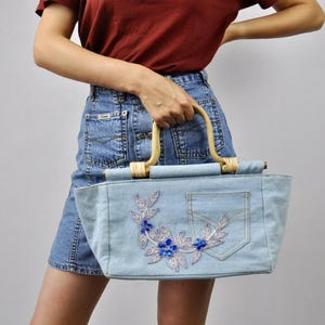 May include: Light blue denim tote bag with a pocket and floral embroidery. The bag has a bamboo handle and is decorated with silver and blue floral embellishments. A stylish and unique handbag.